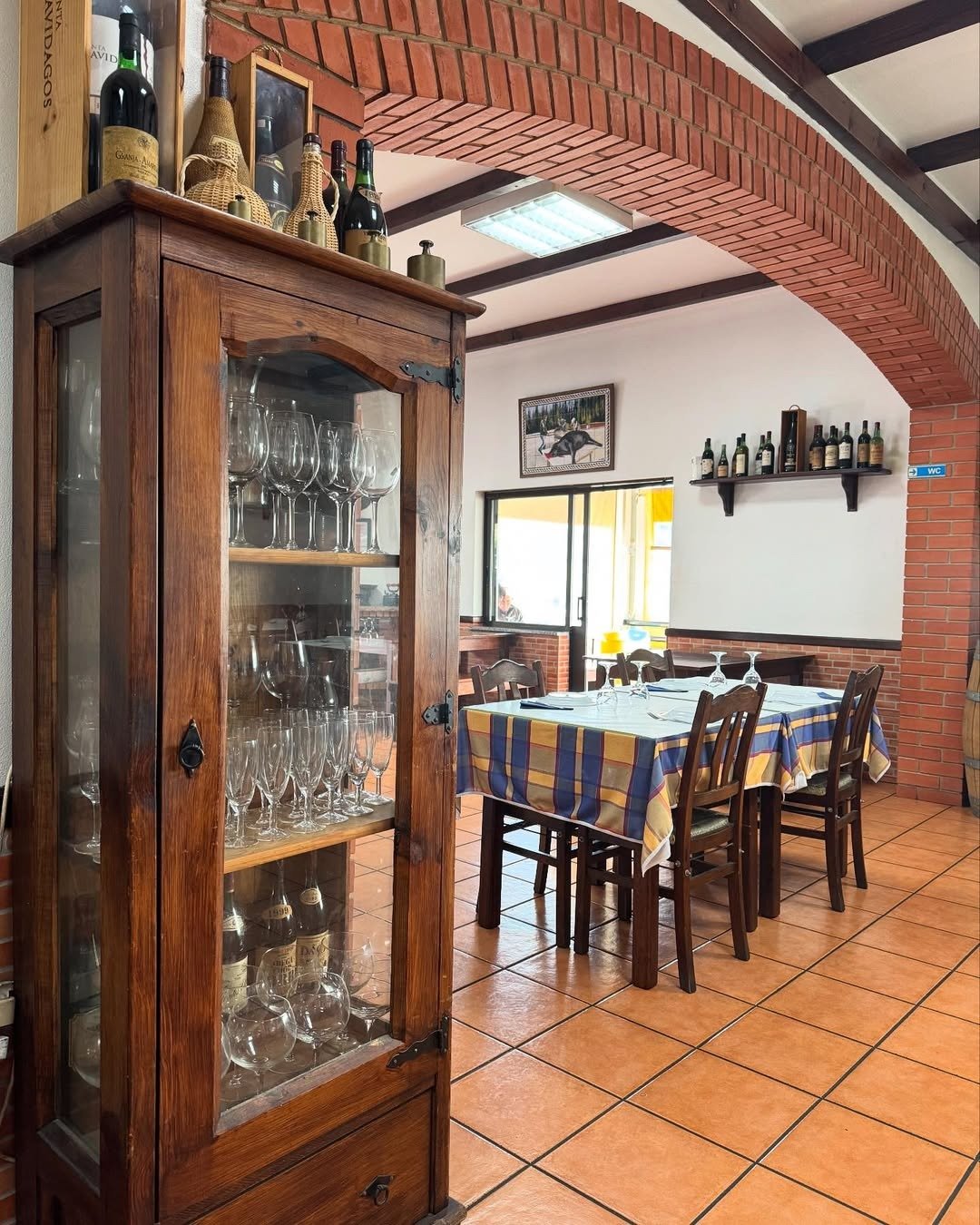 Sala do restaurante com arco de tijolo, vitrine de madeira com copos e garrafas e uma mesa posta com toalha aos quadrados